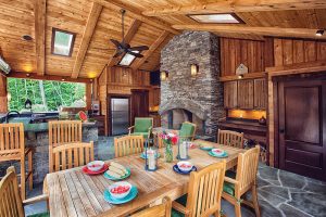 Teak dining table set for 8 people with a fireplace and outdoor kitchen behind it in a log pavilion.